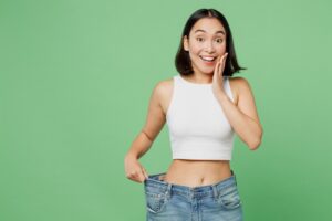 Smiling woman against a green background holding her loose waistband to show the weight she's lost with Phentermine.