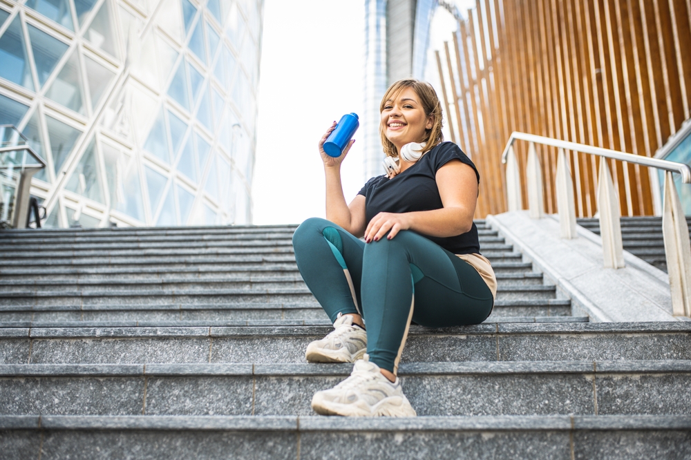 Woman sitting on some steps and drinking her water in the middle of an exercising session.