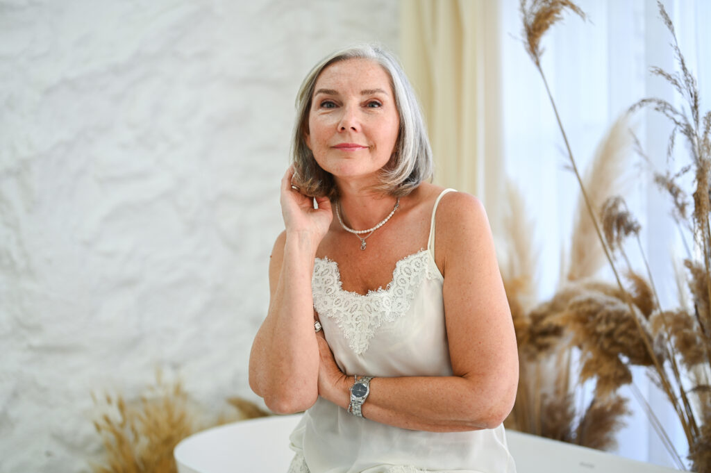 Mature woman with grey hair posing confidently in camisole Mature woman touching her jawline, representing facial contour and volume concerns that may be treated with Radiesse.